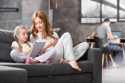 A woman and a girl sitting on a couch looking at a tablet while a man is in the background looking at a computer