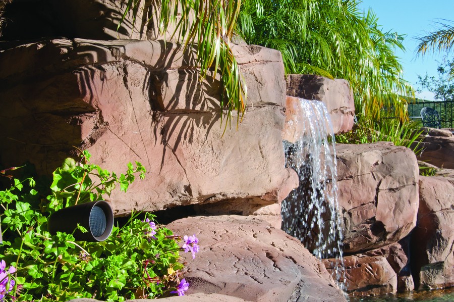 Sonance bullet speaker camouflaged among poolside landscaping featuring greenery and a faux waterfall.