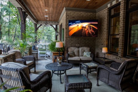 Photo of a brick and concrete patio set up with a plush couch, chairs, a coffee table with a lamp, and an outdoor television.